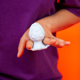 A person in a purple shirt holds a Cute Little Fuckers Princette Puppypus - Platinum, an adorable figurine with large eyes and a tiny mouth, in their hand. The background is a bright orange color.